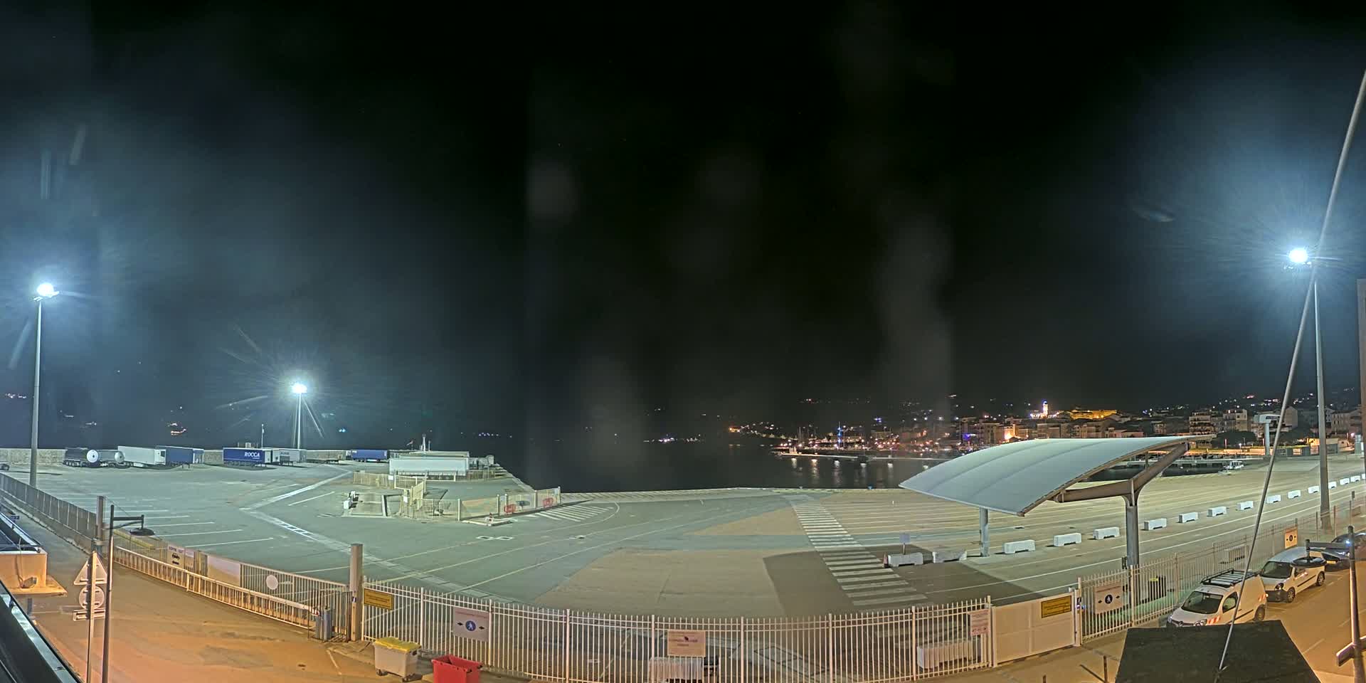 A nighttime view of an empty port area with several parked trucks and a few cars, brightly lit by artificial lights, overlooks a dark body of water with a city skyline in the distance.