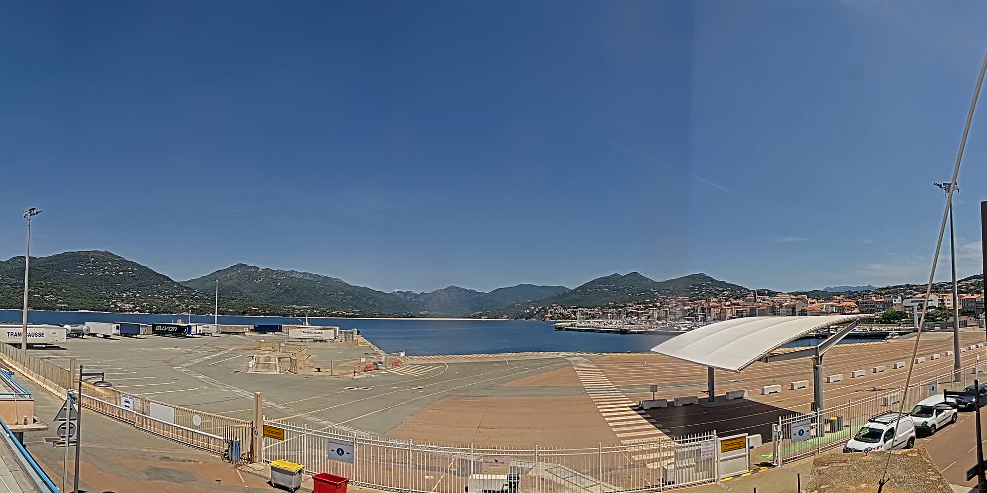 A large, empty paved area next to a calm body of water, surrounded by mountains under a clear blue sky.