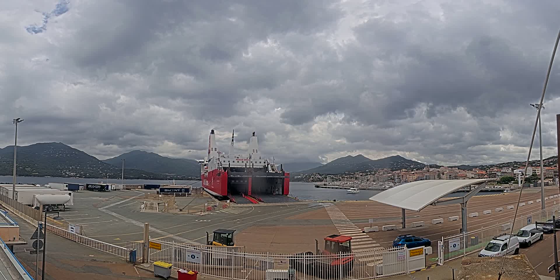 A large red and white ferry is docked at a port under a cloudy sky, with mountains visible in the distance and several vehicles parked nearby.