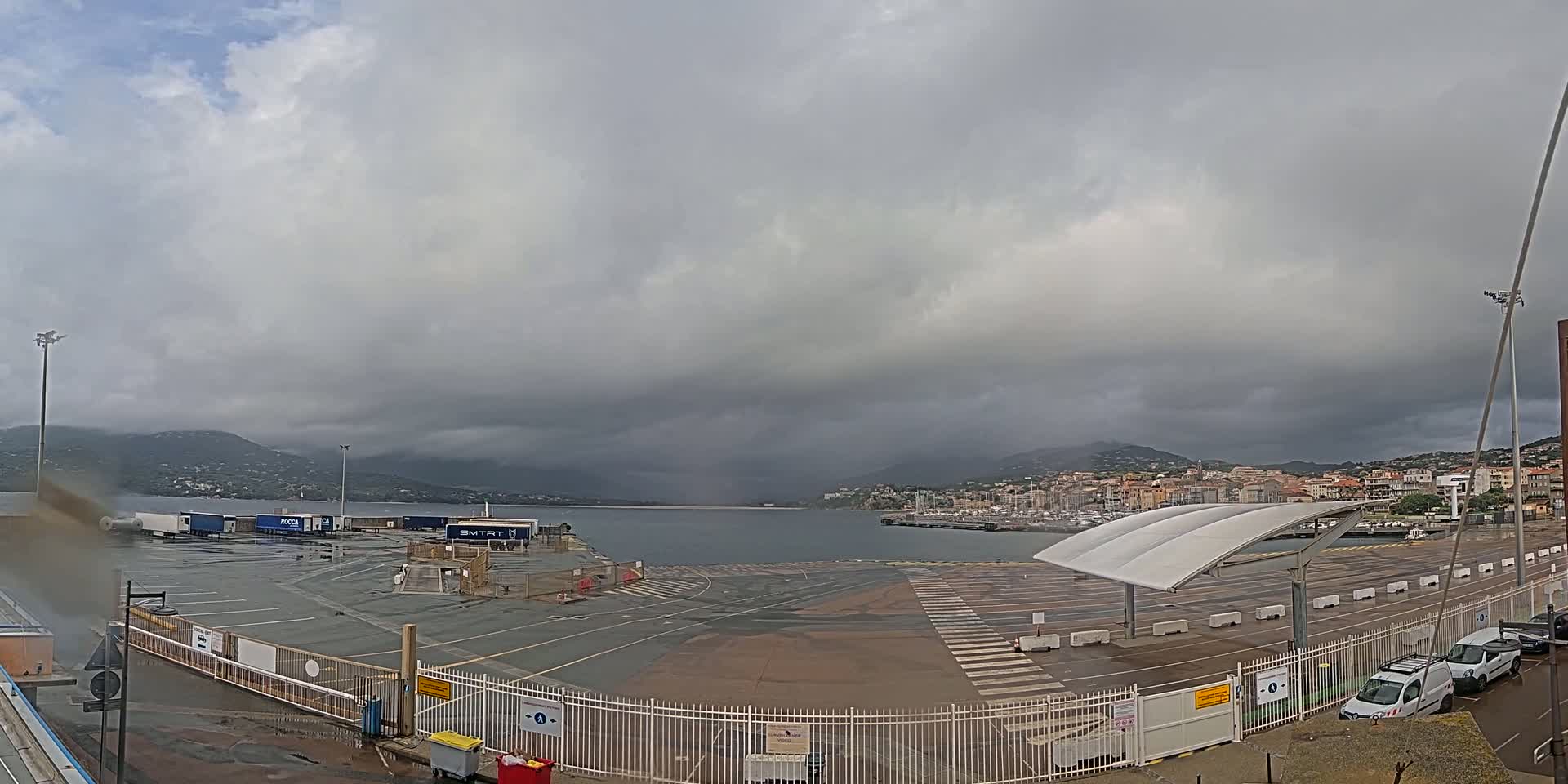 A wet, overcast day at a port, showing a harbor with docked boats, a parking lot with several vehicles, and cargo trucks in the foreground.
