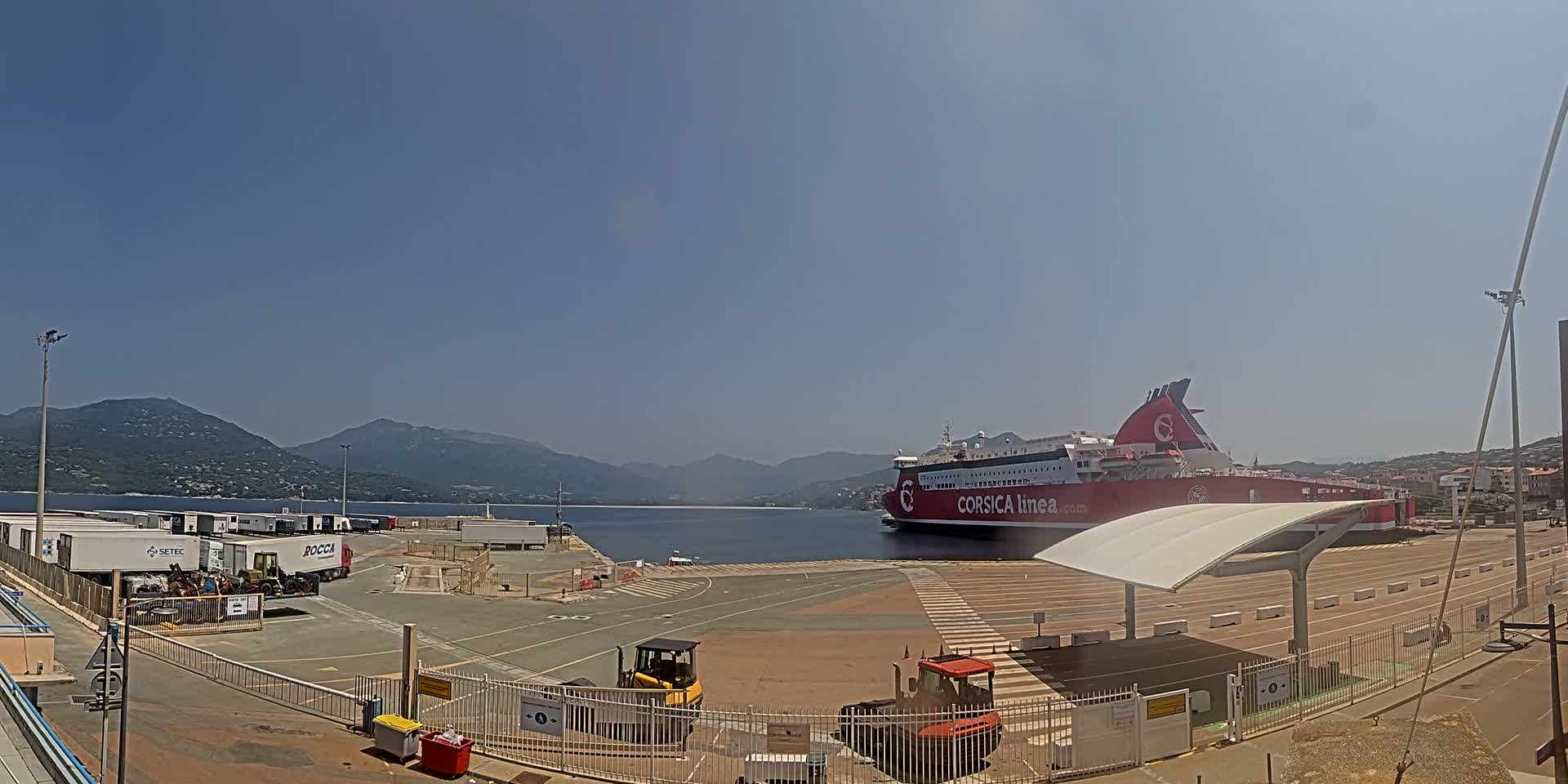 A large red and white ferry is docked at a port under a clear, sunny sky, with mountains in the background and several pieces of heavy equipment in the foreground.