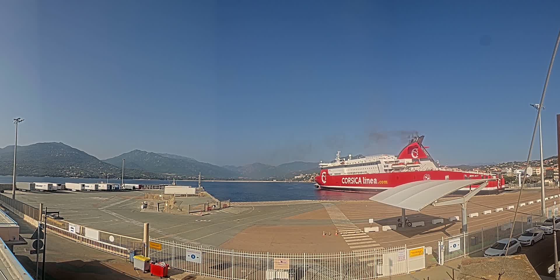 A large red ferry is docked at a port on a clear, sunny day, with mountains visible in the background.