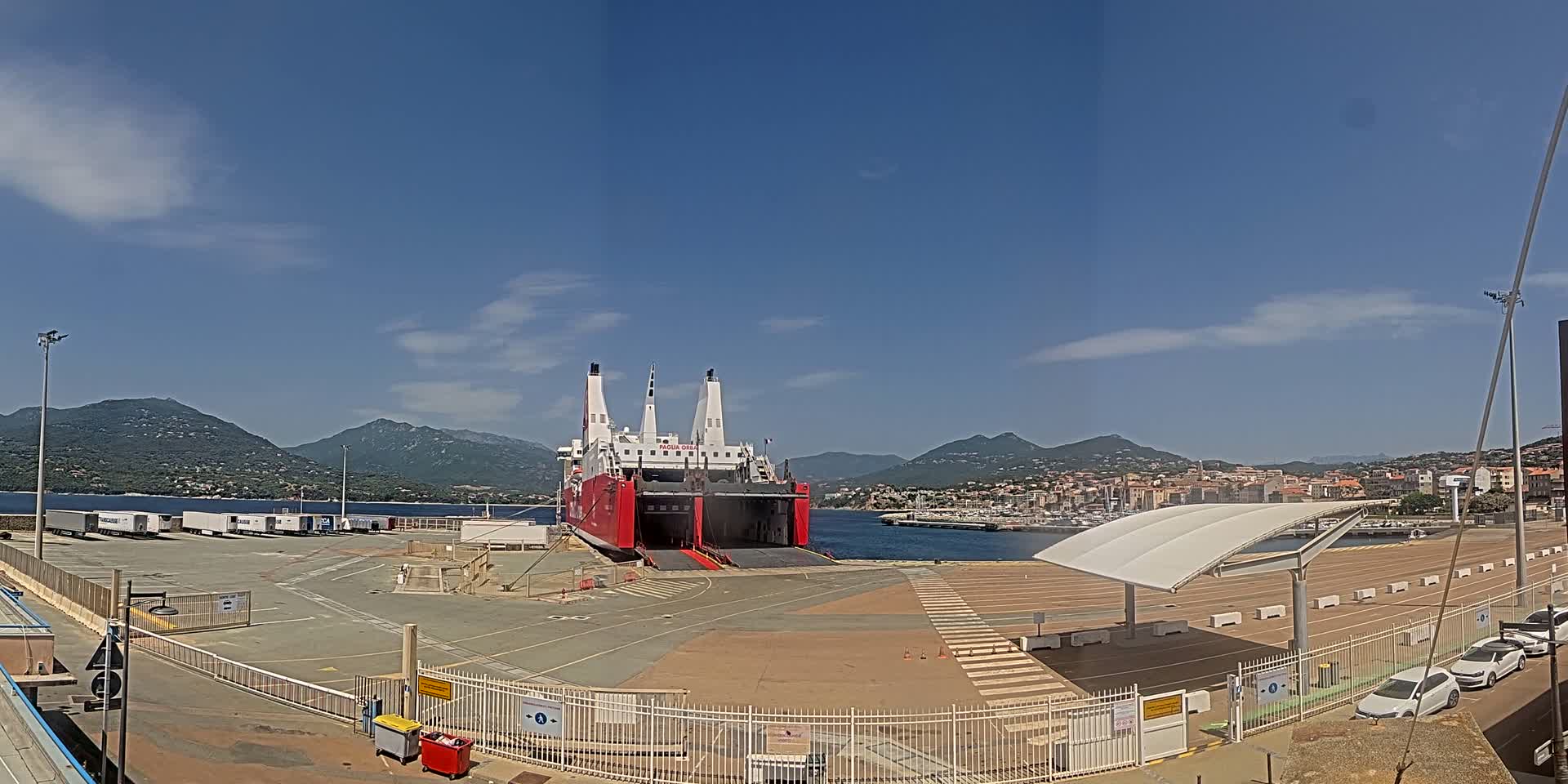A large red and white ferry is docked at a port, with several trucks and cars parked nearby, under a mostly clear blue sky and mountains in the distance.