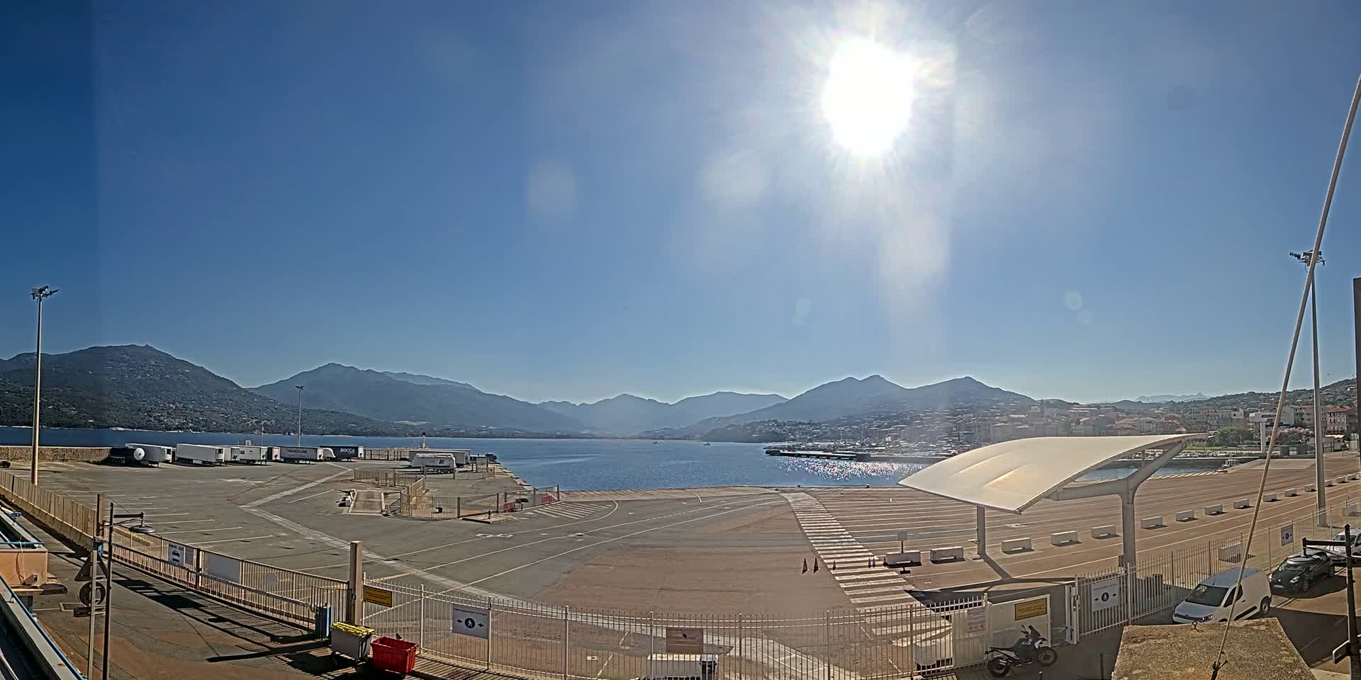 A sunny day shows a harbor with several trucks parked in a lot, a calm body of water, and mountains in the distance.