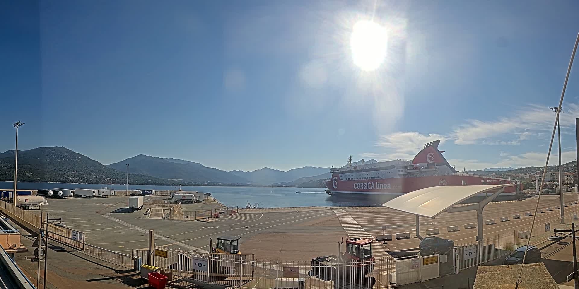 A large red ferry is docked at a port on a sunny day with mountains visible in the distance.
