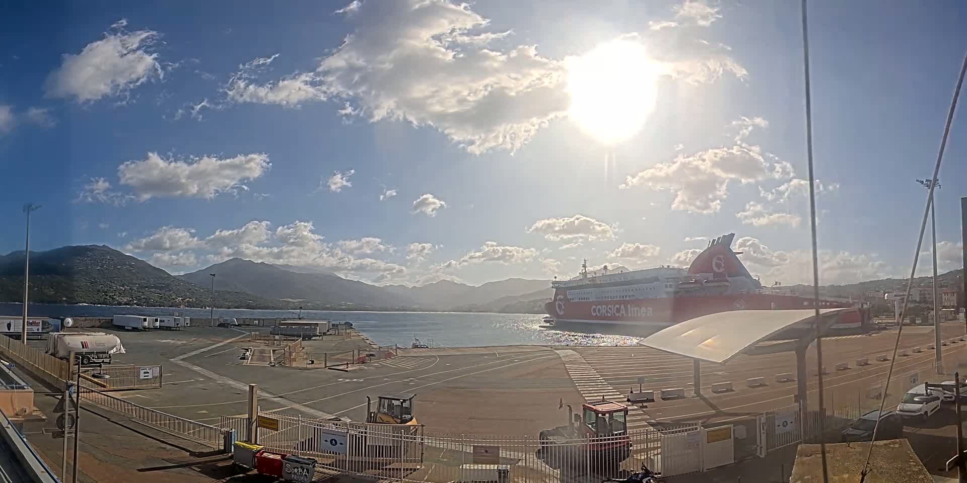 A large ferry is docked at a port under a mostly sunny sky with scattered clouds and mountains in the distance.