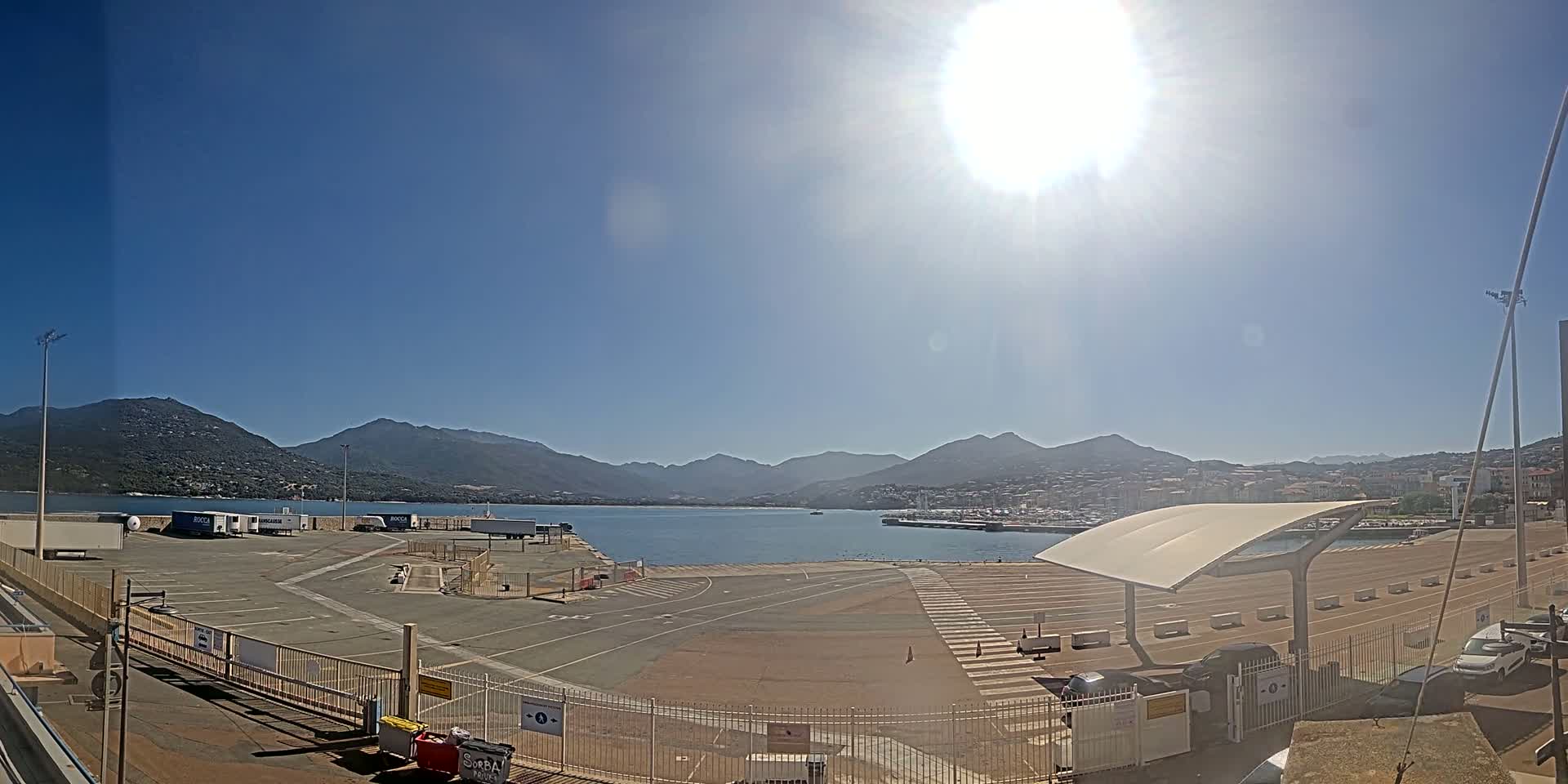 A sunny day reveals a harbor area with a mostly empty parking lot, a few vehicles, and mountains in the background.
