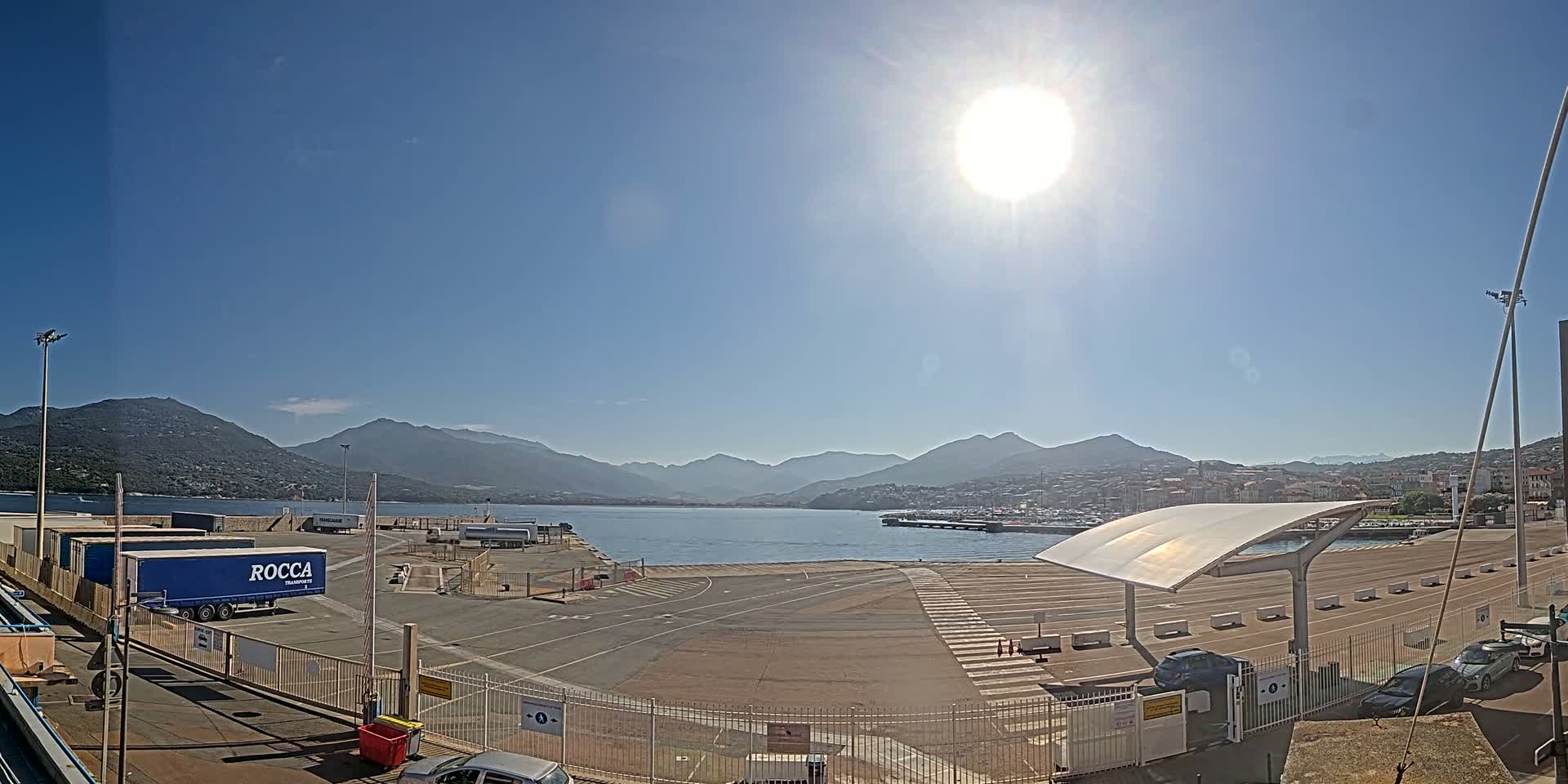 A sunny day at a port, featuring a calm body of water, mountains in the distance, and a semi-empty parking lot with several vehicles.