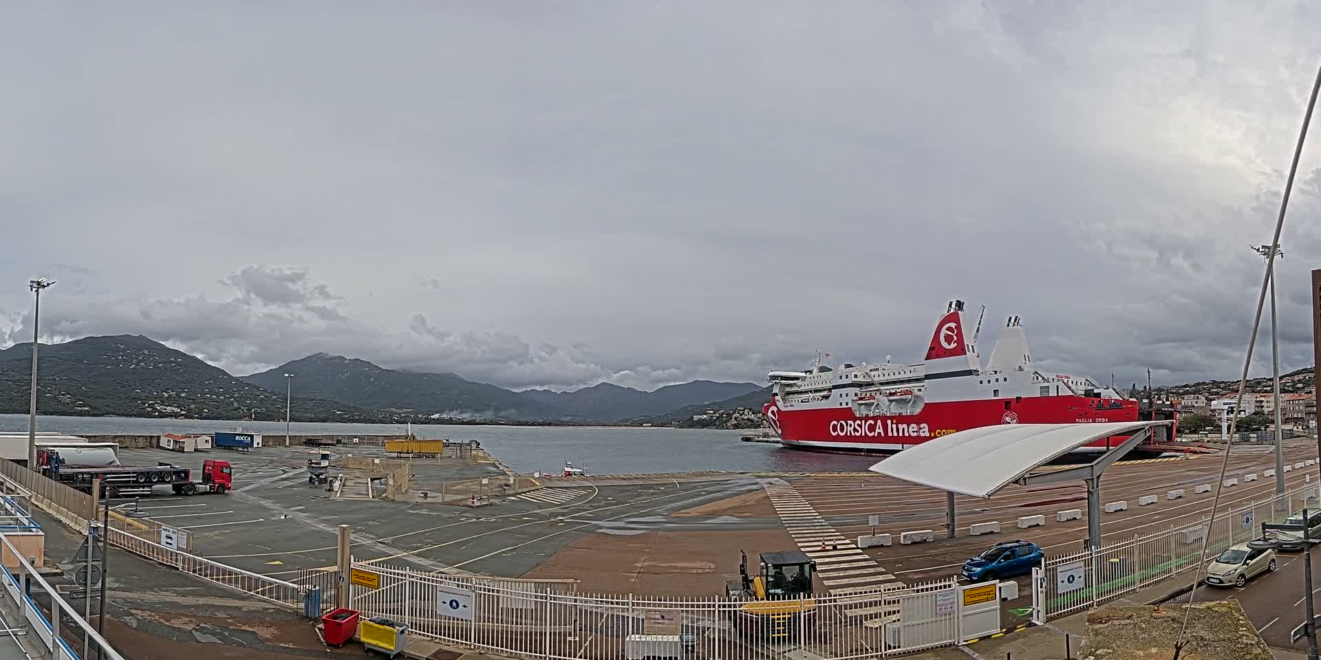 A large red and white ferry is docked at a busy port under a cloudy sky, with several trucks, cars, and construction equipment visible in the foreground and verdant mountains rising from the water in the distance.
