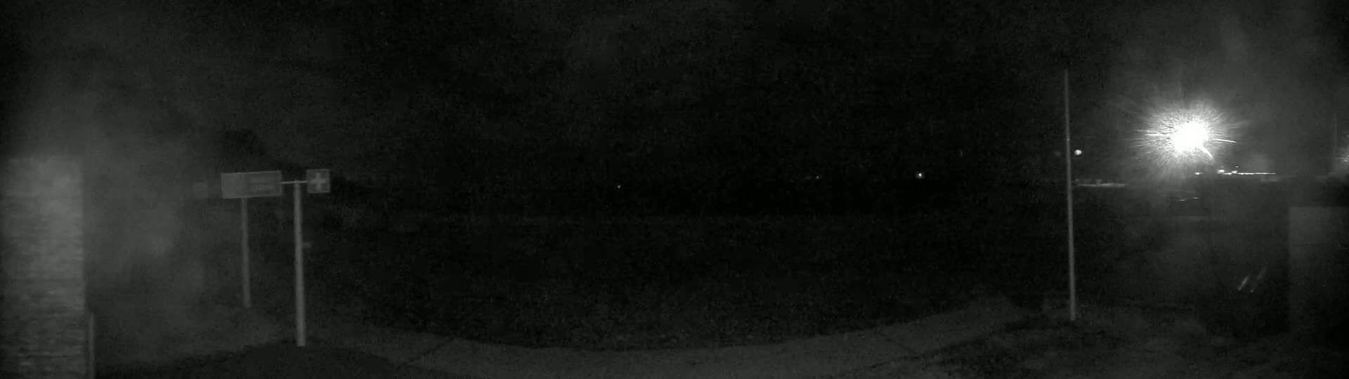 A nighttime, low-light image shows a dark, empty roadway with indistinct objects and a bright light source in the distance.