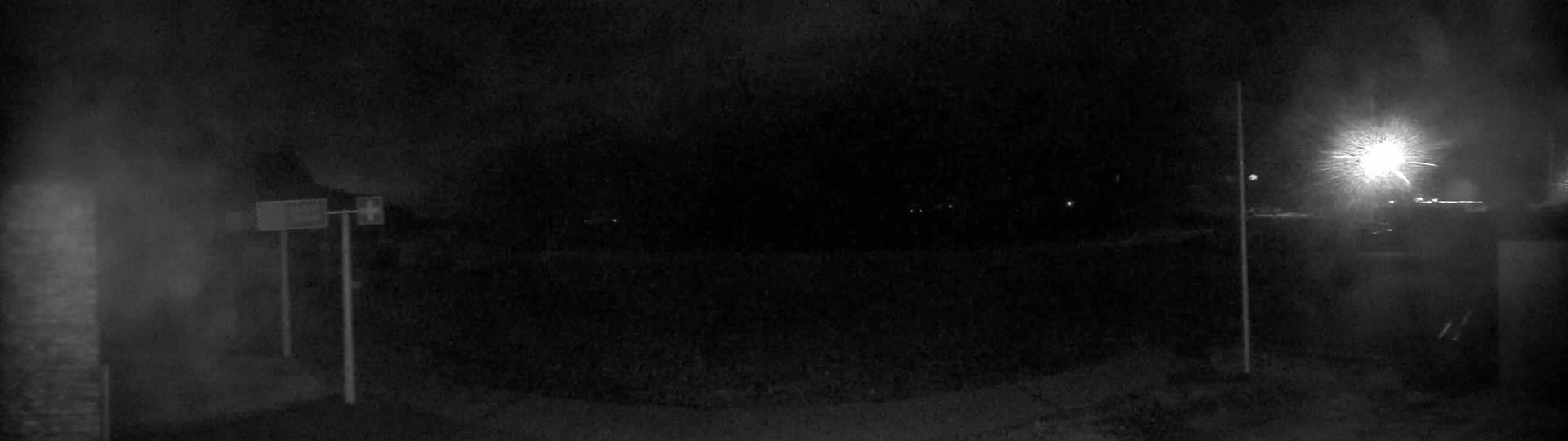 A nighttime, low-light view of a dark, mostly empty area with a bright light source in the upper right corner and two posts with signs in the left.