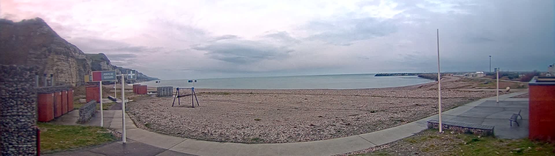 A mostly cloudy day shows a pebble beach with a walkway, a few small buildings, and a calm sea, backed by high cliffs.