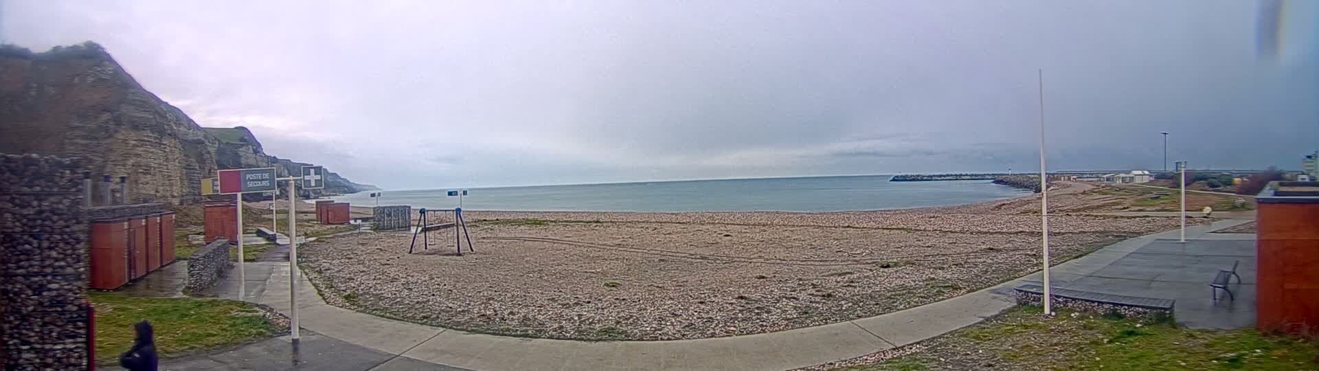 A mostly empty pebble beach stretches to a calm sea under an overcast sky, backed by cliffs and a few small buildings.