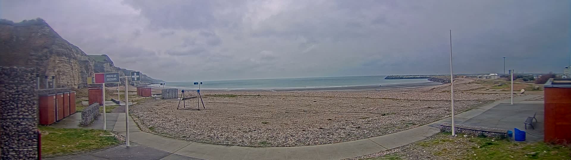 A pebble beach stretches out to a calm, gray sea under a cloudy sky, backed by a tall, rocky cliff and a low sea wall.