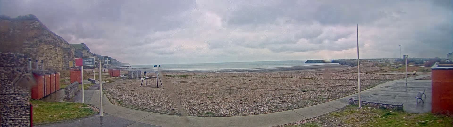 A mostly empty pebble beach is seen under an overcast sky, with a tall cliff to the left and a low sea wall to the right.
