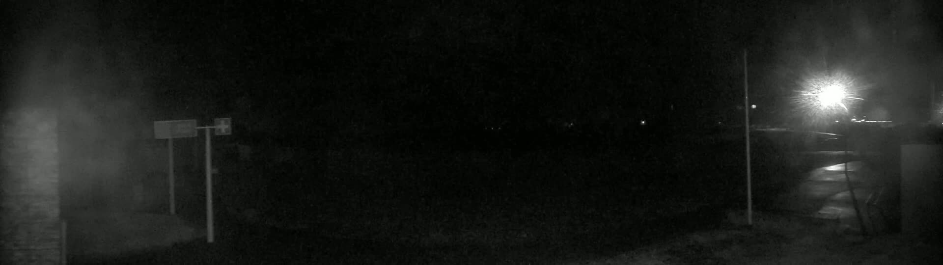 A dark, snowy night scene shows two posts with signs, a bright light in the distance, and a dimly lit area in the foreground.