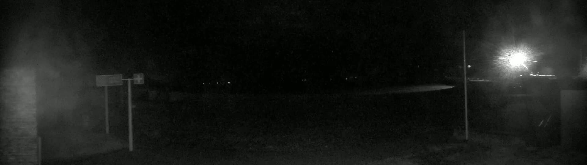 A nighttime, dimly lit scene shows a dark, empty road with a bright light source in the distance and two tall posts with signs.