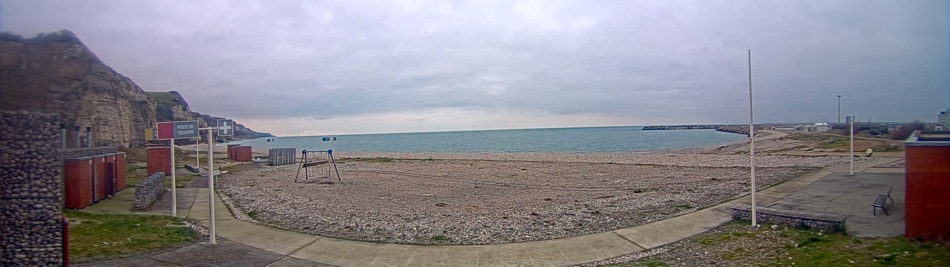 A pebble beach with a small playground and a calm, grey sea under an overcast sky is visible, backed by a tall, light-brown cliff face.