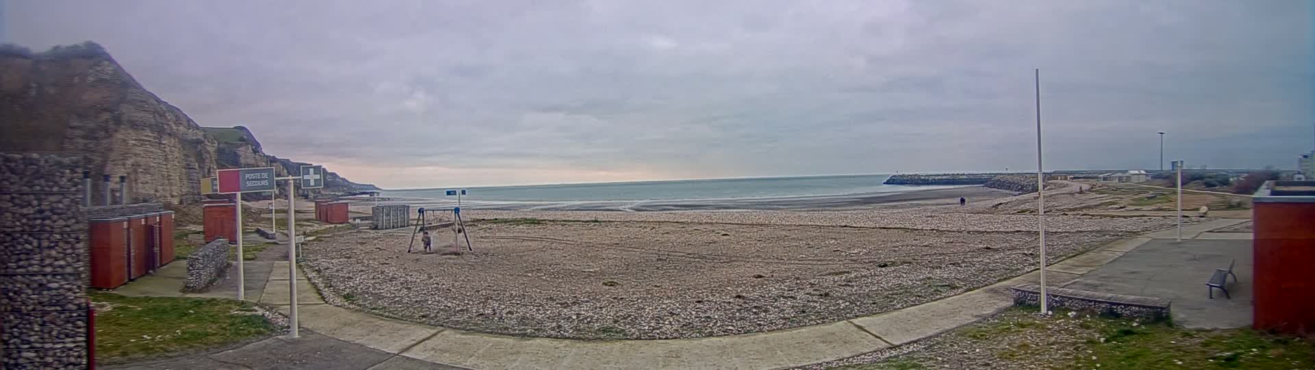 A mostly cloudy day reveals a pebble beach with a walkway, small buildings, and a distant cliff overlooking a calm sea.