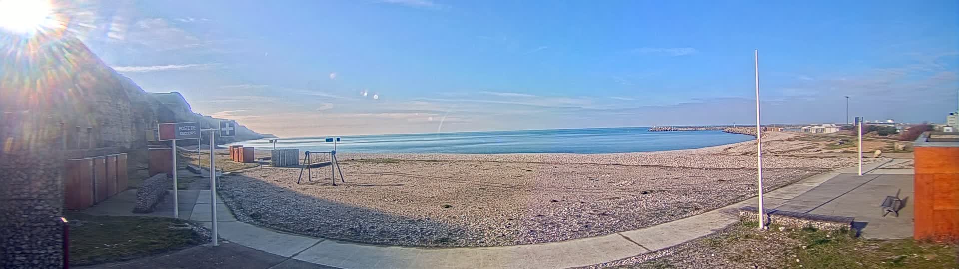 A sunny day at a beach with calm blue water, a sandy shore, a small playground, and a rocky cliff in the background.