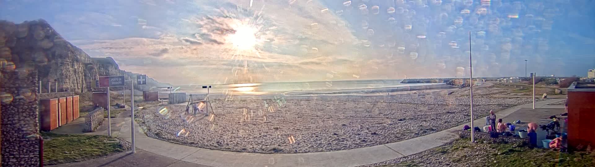A mostly sunny beach scene with a few people near the shore and buildings, viewed through a rain-covered window, shows a rocky cliff face to the left.
