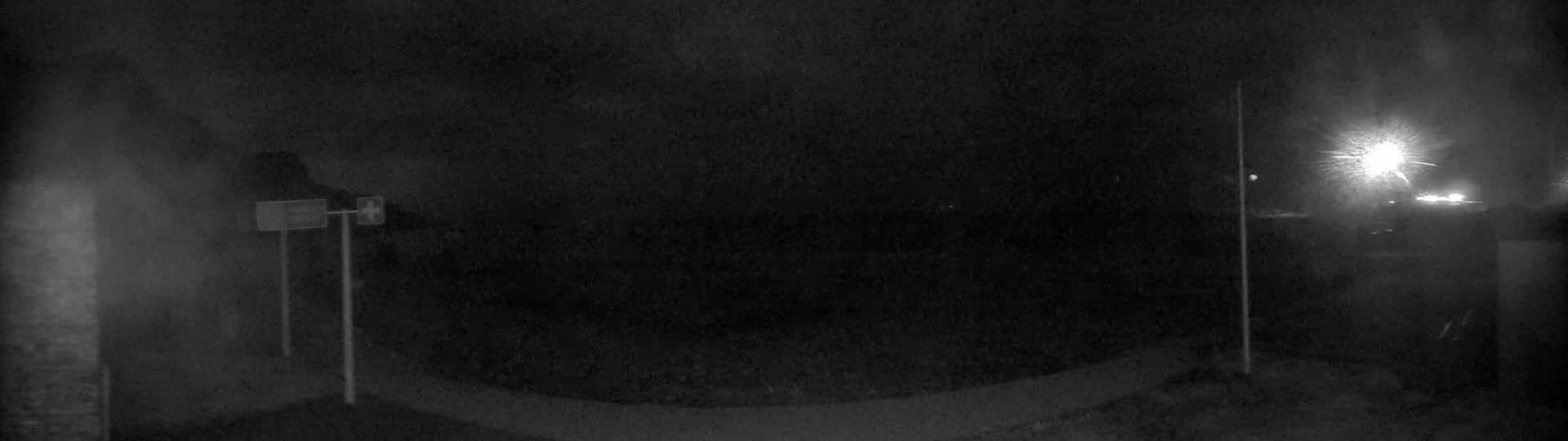 A nighttime, low-light image shows a dark, mostly empty area with two tall posts and a curved surface in the foreground, and bright lights visible in the distance.