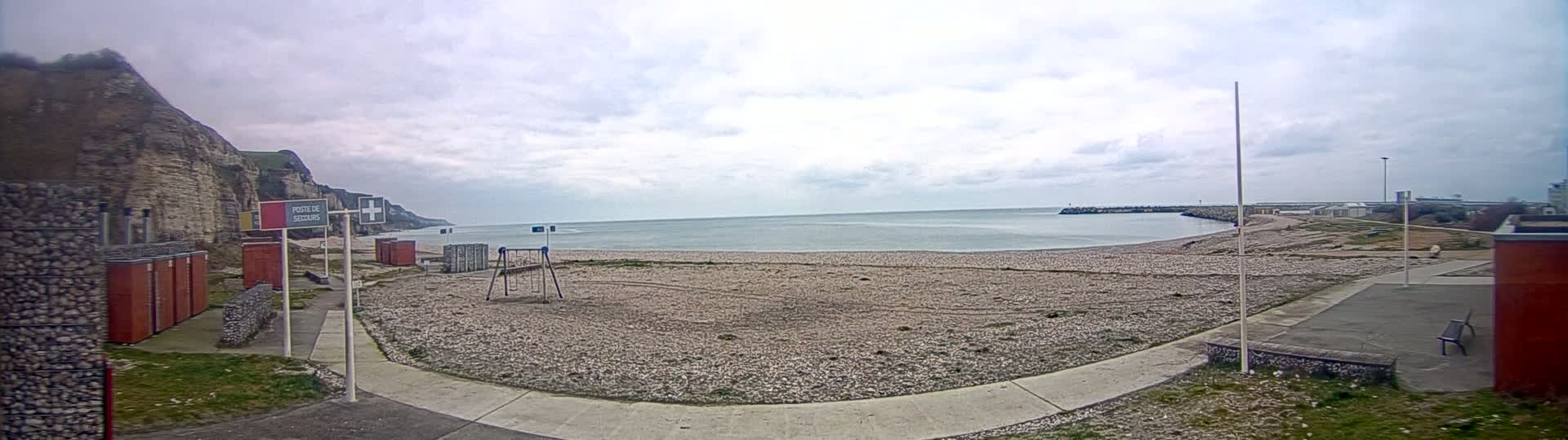 A mostly empty pebble beach is visible under an overcast sky, with a playground, a small breakwater, and white cliffs in the background.