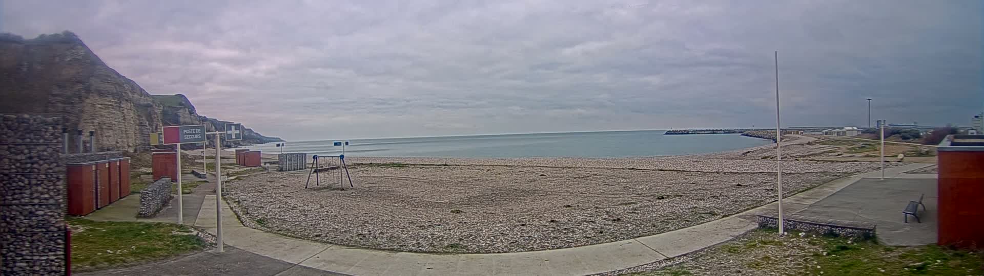 A pebble beach stretches out to a calm sea under an overcast sky, backed by a rocky cliff face and some small buildings.