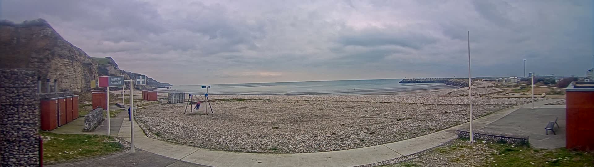 A mostly empty pebble beach stretches towards a calm, gray sea under a cloudy sky, with a few buildings and a swing set visible nearby.