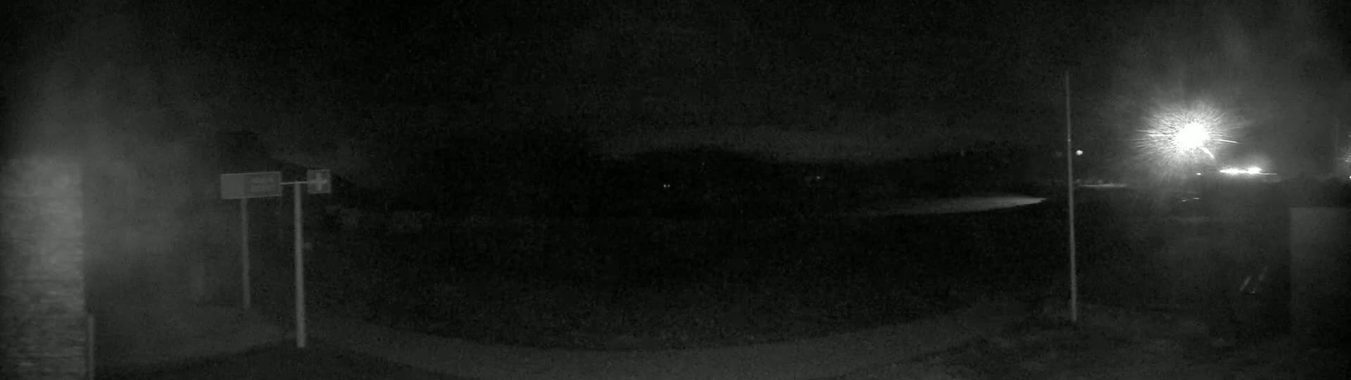 A nighttime, low-light view shows a dark, empty area with some faint lights in the distance and two upright poles near the bottom left.