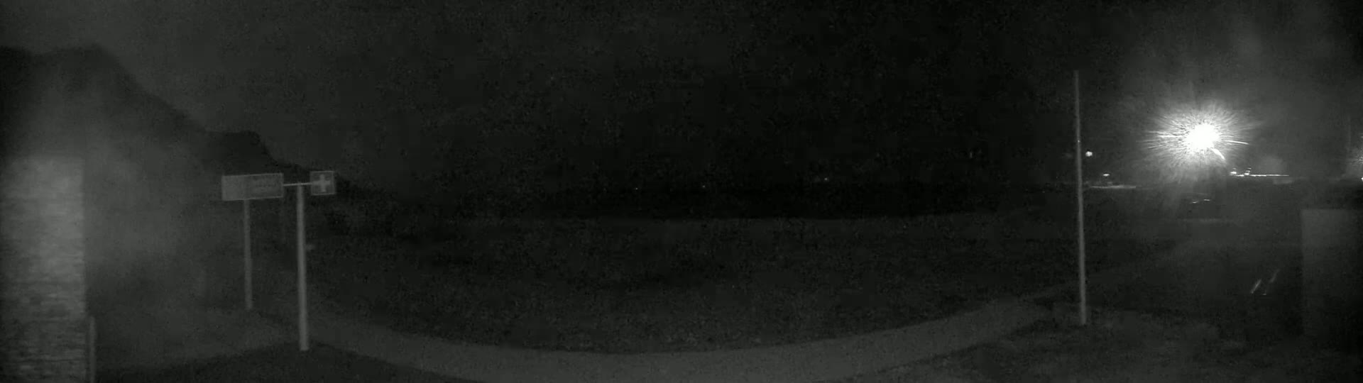 A nighttime, low-light image shows a paved area with two poles and signs, a blurry background, and a bright light source in the upper right corner.