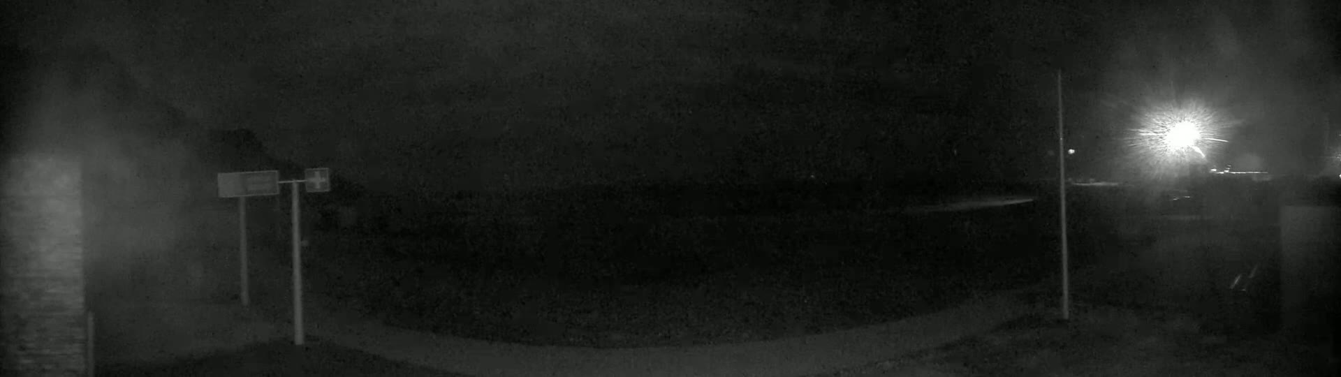 A nighttime, low-light image shows a dark paved area with two poles, possibly light poles, and a bright light source in the distance.