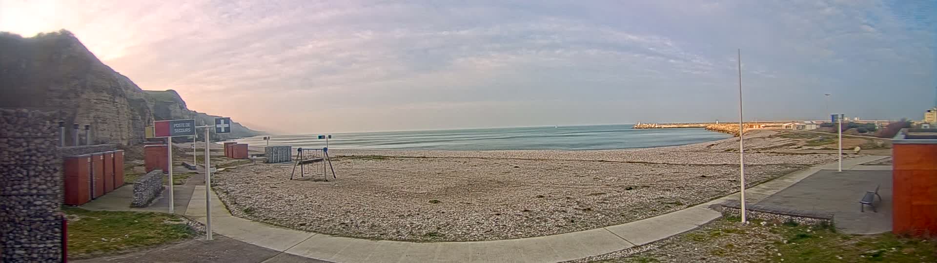 A mostly empty pebble beach stretches between a rocky cliff and a harbor under a partly cloudy sky.