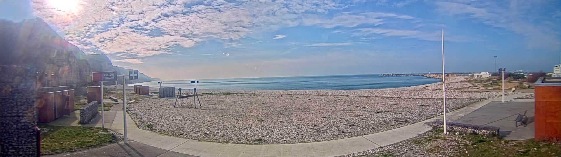 A mostly sunny day reveals a pebble beach with calm ocean water, a small playground, and a rocky cliff in the background.