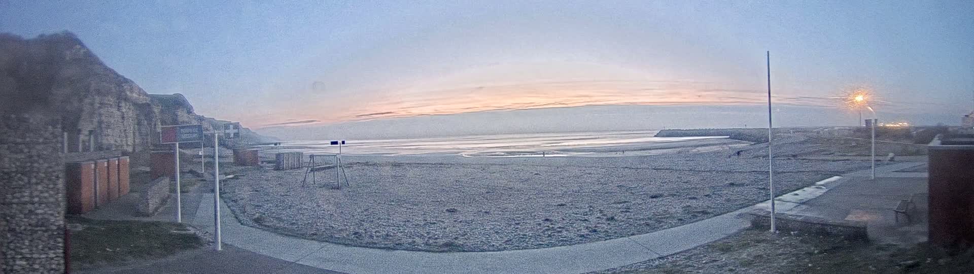 A mostly empty pebble beach at sunset is visible from a vantage point near a stone building, with calm ocean water in the background.