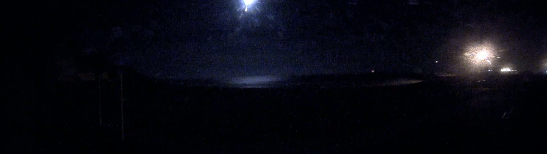 A dark, nighttime scene shows a bright light in the distance over a dark body of water, illuminated by other distant lights.