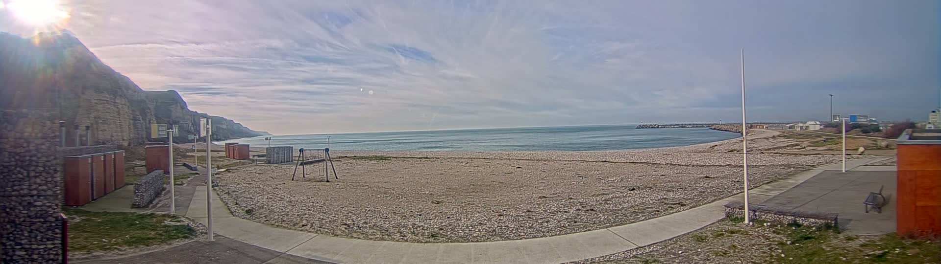 A mostly sunny day at a pebble beach with a calm sea, a playground, and a cliff in the background.