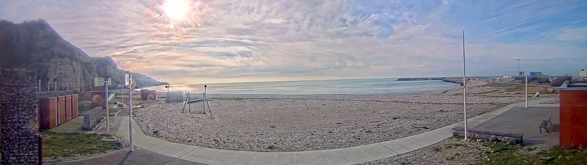 A mostly sunny day reveals a pebble beach with a calm sea, a cliff face to the left, and a breakwater to the right.
