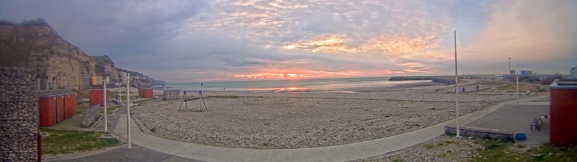A pebble beach at sunset, under a partly cloudy sky, is seen from a vantage point near a playground and changing rooms nestled against a cliff face.