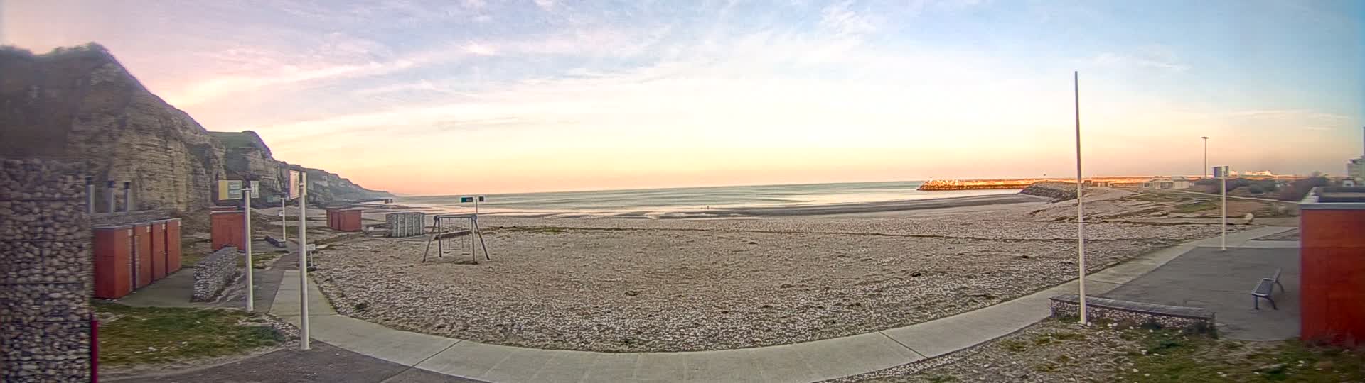 A pebble beach at sunrise, with a low sea wall and white cliffs visible in the background, under a mostly clear sky.