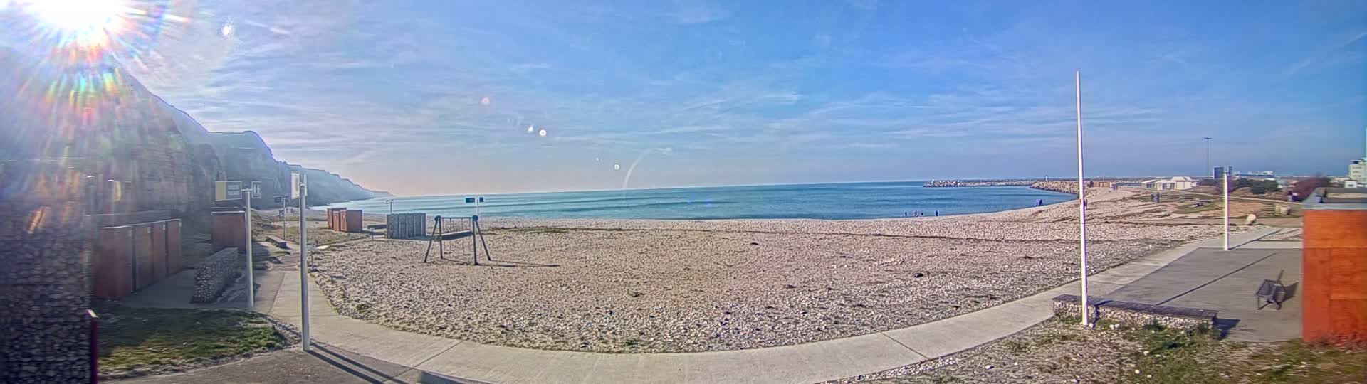 A sunny day at a pebble beach with a calm sea, a playground, and a cliff in the background.