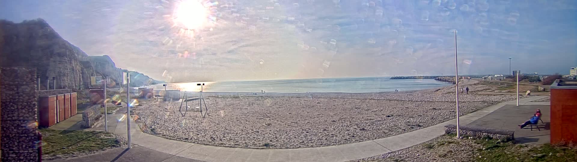 A sunny day at a pebble beach shows a calm sea, a cliff in the background, and a person sitting on a bench near a building.