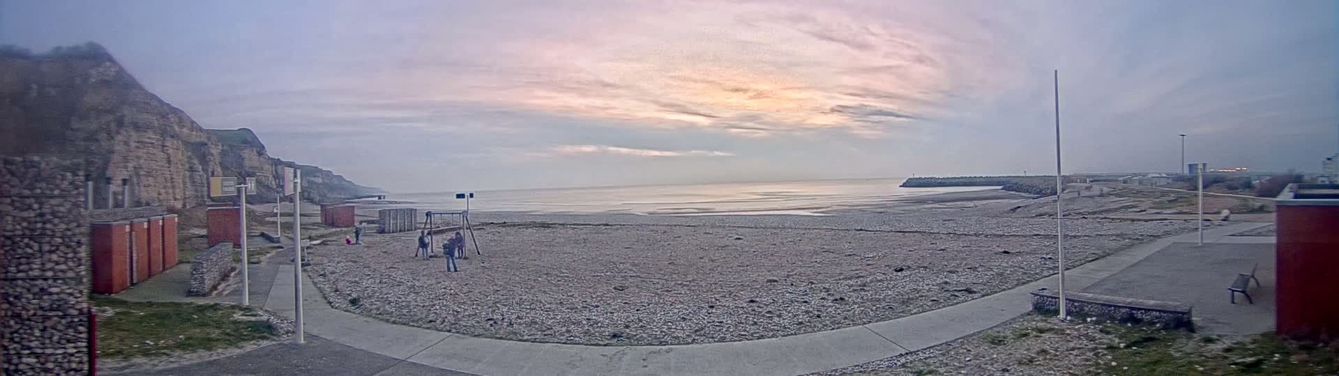 A pebble beach stretches out to a calm sea under a pale, cloudy sky with hints of pink in the horizon.