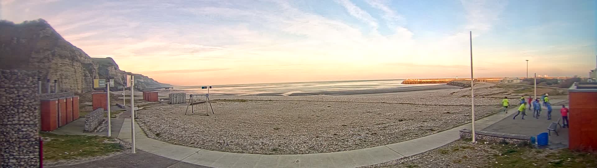 A group of people are gathered on a pebble beach at sunset near a rocky cliff, with calm ocean waters extending to the horizon.