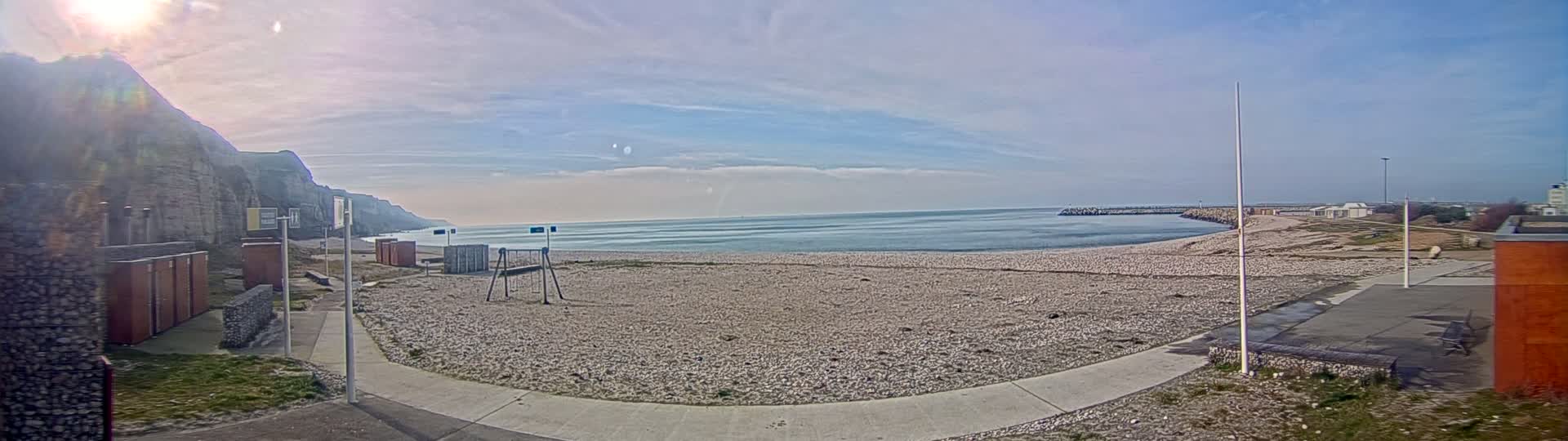A mostly sunny day at a pebble beach with a small playground, overlooking calm ocean water and a rocky cliff.