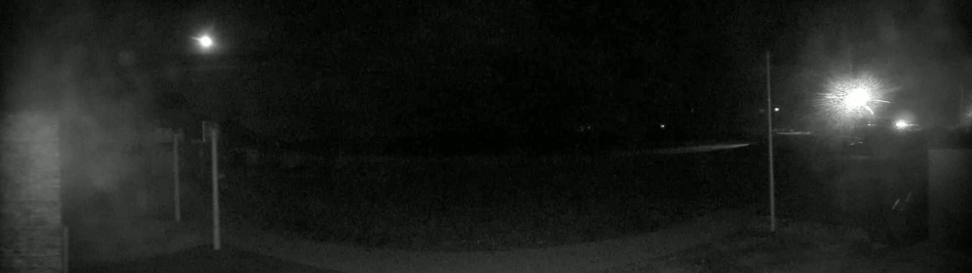 A nighttime, low-light outdoor scene shows a dark, paved area illuminated by several distant lights under a mostly clear sky.