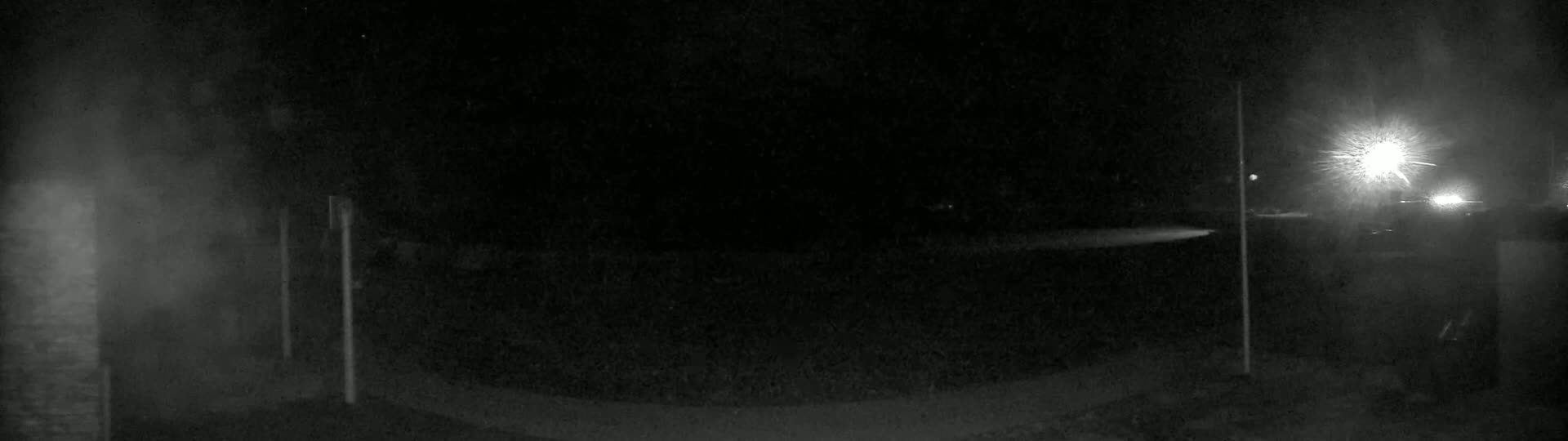 A dark, nighttime scene shows a paved area illuminated by a bright light source in the distance, with vertical posts visible on either side.