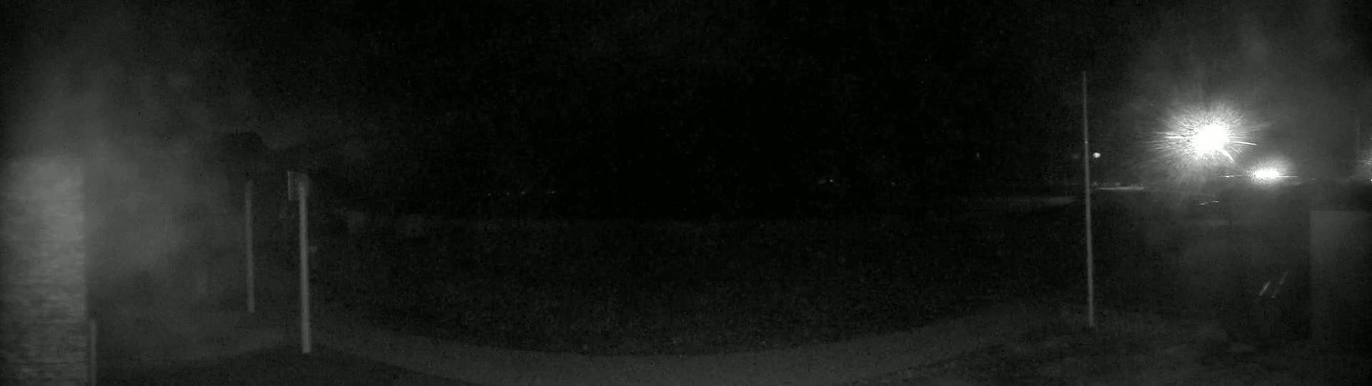 A dark, nighttime scene shows a paved area with two tall, thin poles and bright lights in the distance.