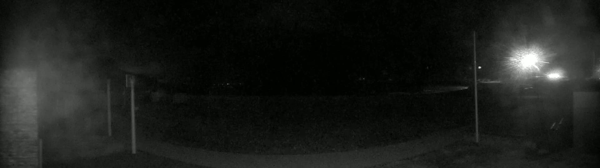 A nighttime, low-light view of a dark, paved area with two vertical poles and blurry lights in the distance.