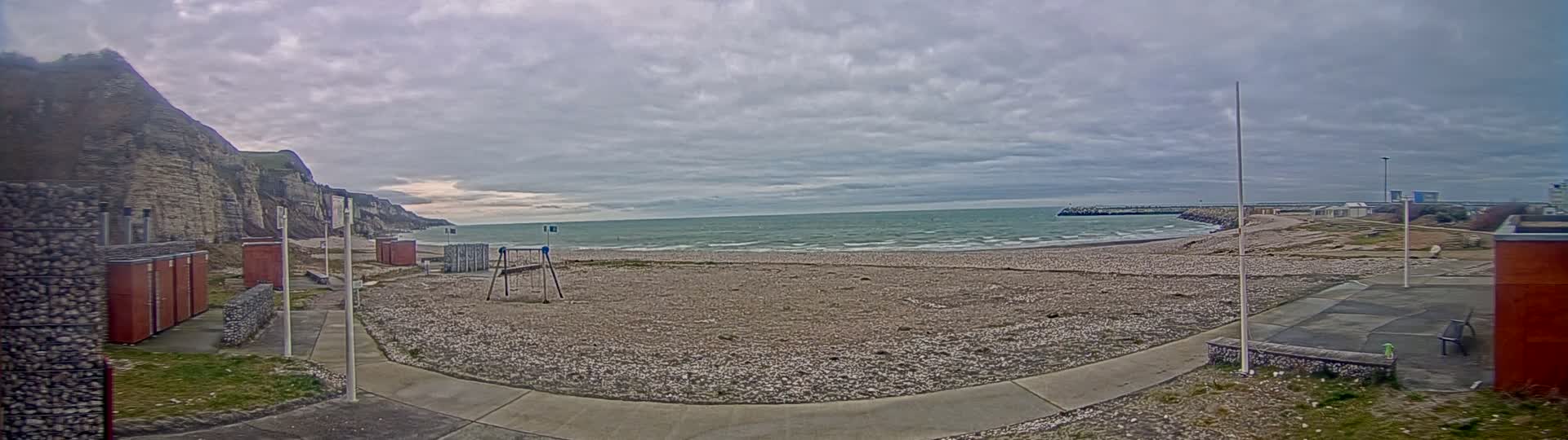 A mostly empty pebble beach stretches between a rocky cliff and a harbor entrance under a cloudy sky, with a small playground and changing rooms visible.